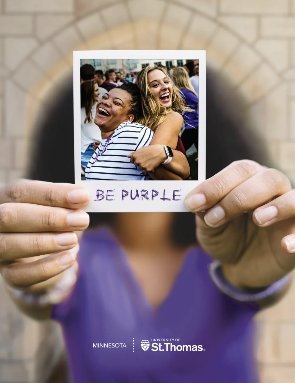 A woman in purple holding a polaroid picture of two Tommies smiling with Be Purple as the polaroid caption