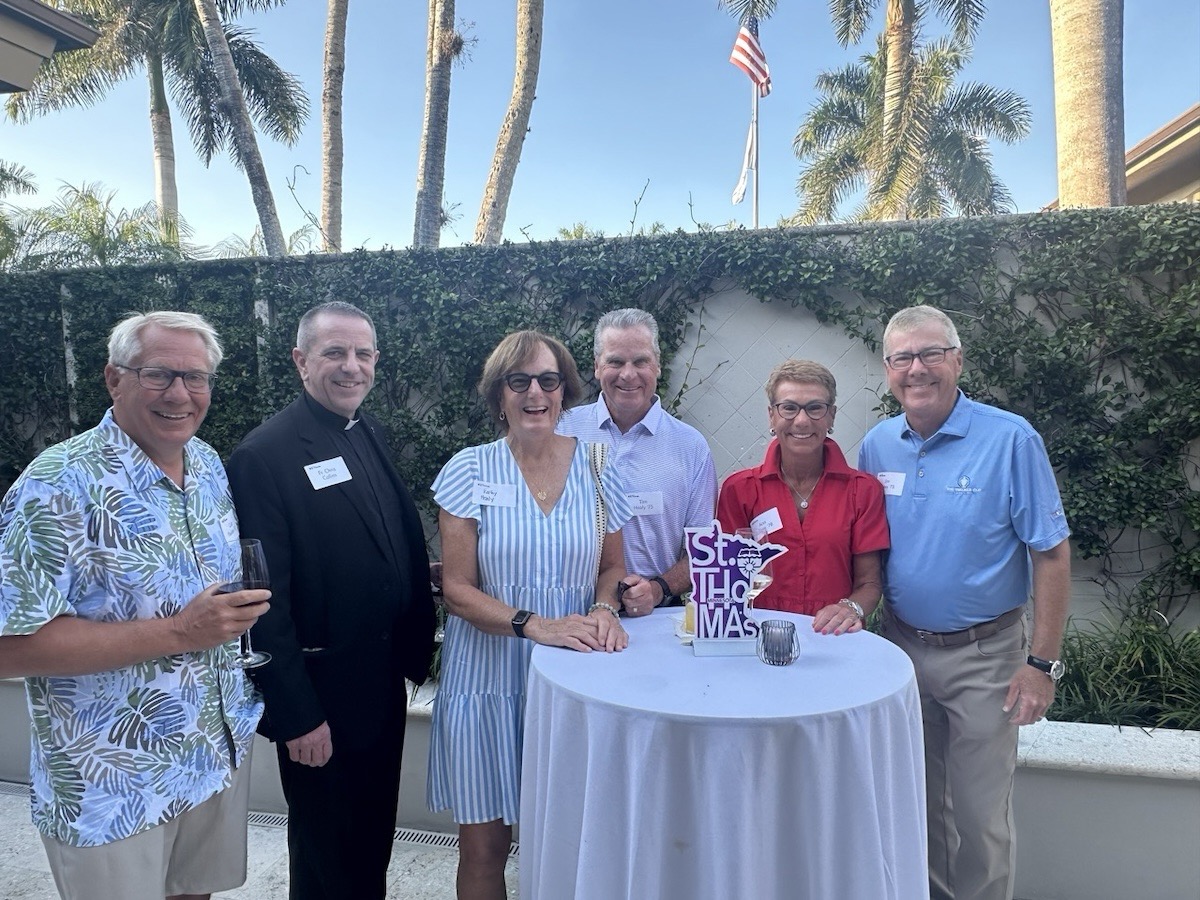 Father Chris poses for a photo with five Tommie alumni. They're smiling and holding drink glasses.