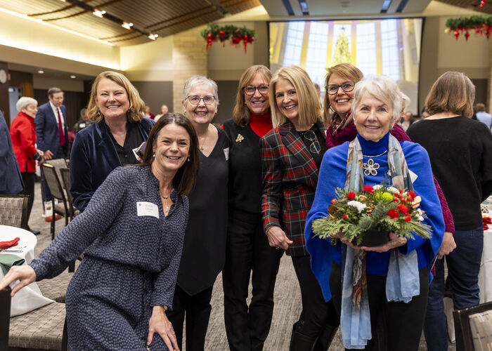 Attendees pose for a photo at the Alumni and Friends Christmas Luncheon in Woulfe Alumni Hall on December 6, 2024 in St. Paul. They’re wearing festive holiday clothes and holding Christmas flower arrangements.