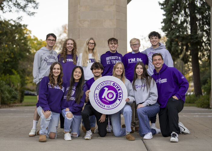 A group of 12 Tommie students wear St. Thomas gear and pose with a Tommie Give Day sign in front of the arches.