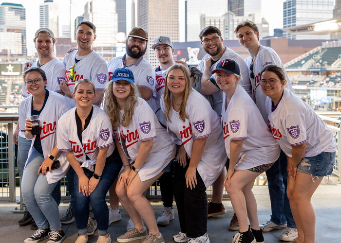 12 Tommie alumni pose for a photo at Target Field for St. Thomas Night with the Twins 2024. They’re wearing matching jerseys that have Twins and St. Thomas logos.