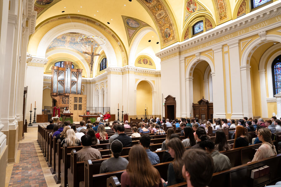 The interior of the Chapel of St. Thomas Aquinas during mass. The pews are full of worshipers and a woman is speaking at the pulpit.