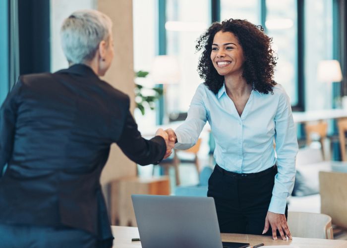 Two people in professional dress shake hands over a desk.