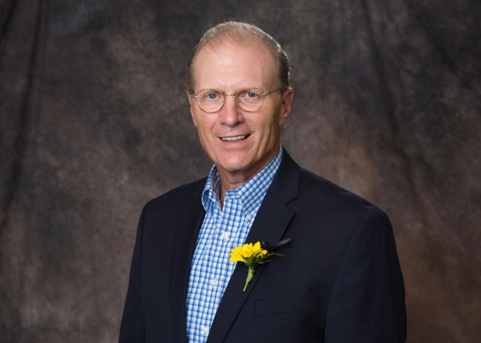 Mark C. Dienhart, PhD ’75 smiles into the camera for a studio portrait taken against a brown backdrop