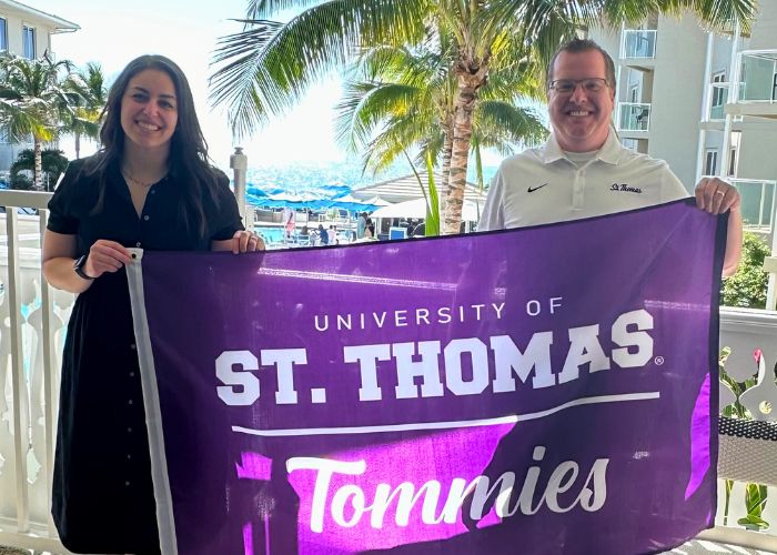 Tierney Wilson and Ben Fraser smile for the camera while holding a purple flag that reads "University of St. Thomas Tommies"