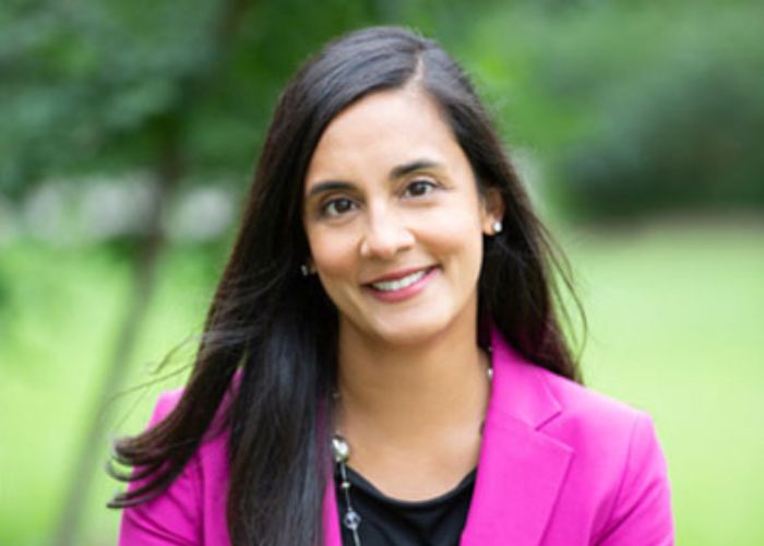 Ulcca Joshi Hansen wears a pink blazer and smiles for a portrait in front of a lush green outdoor background