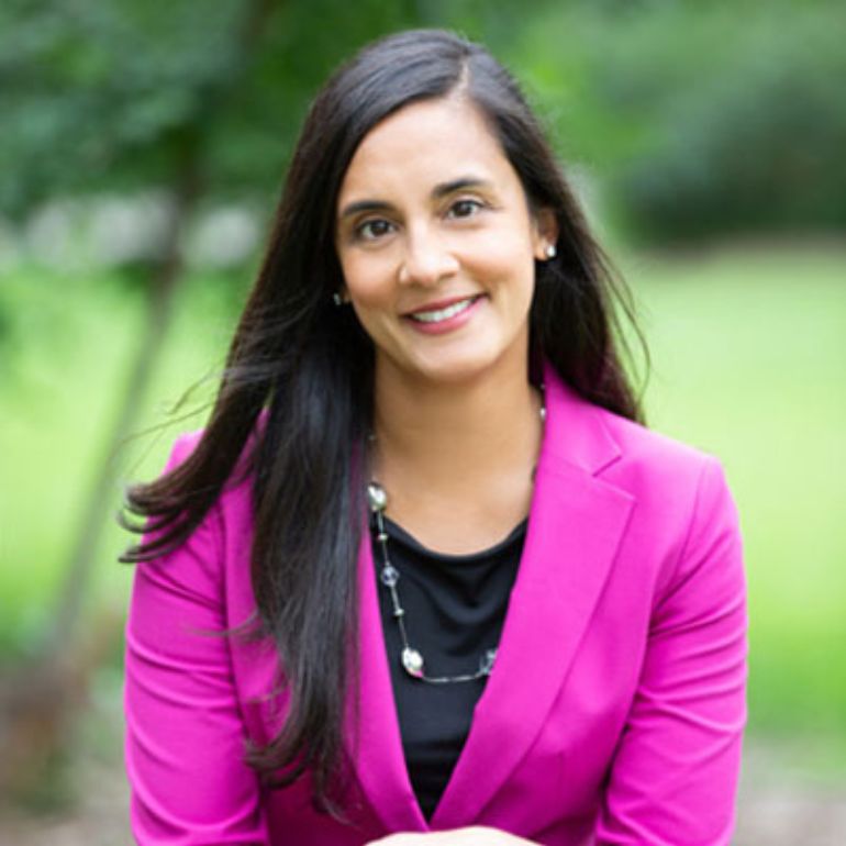 Ulcca Joshi Hansen wears a pink blazer and smiles for a portrait in front of a lush green outdoor background