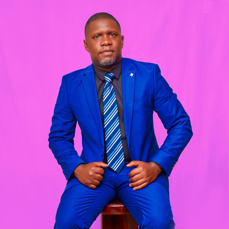 Charles Morgan Kisitu wears a blue suit and poses for a studio portrait in front of a bright pink background