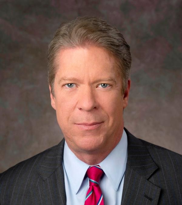 Major Garrett looks into the camera for a studio portrait taken in front of a brown background.