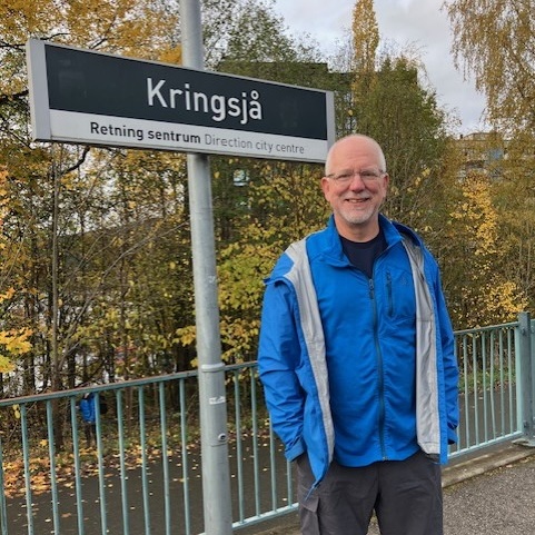Larry wears a blue jacket and stands smiling in front of a train station sign