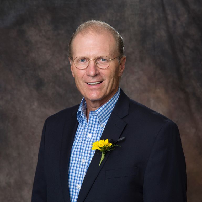 Mark C. Dienhart, PhD ’75 smiles into the camera for a studio portrait taken against a brown backdrop